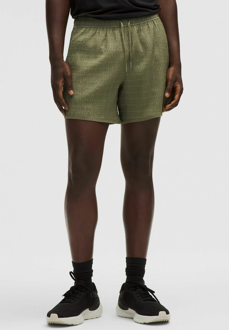 Olīvzaļi sporta šorti ar teksturētu virsmu, elastīgu jostu un auklu. Apvienoti ar melniem sporta apaviem un zeķēm.