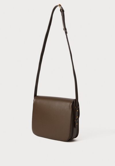 Sac en cuir marron foncé à bandoulière réglable et fermeture à rabat, présenté suspendu sur un fond clair et uni.