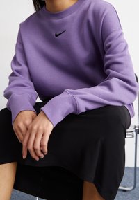 Iemand zit en draagt een paarse Nike-sweater en een zwarte rok, met de handen op de schoot en de vingertoppen die lichtjes elkaar raken.