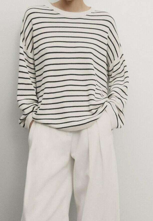 Personne portant un pull blanc ample avec de fines rayures horizontales noires et un pantalon blanc large à taille haute, mains dans les poches.