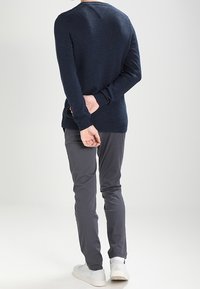 Person steht mit dem Rücken zur Kamera, die Hände hinter dem Rücken gefaltet, trägt einen marineblauen Pullover, graue Hosen und weiße Sneakers.