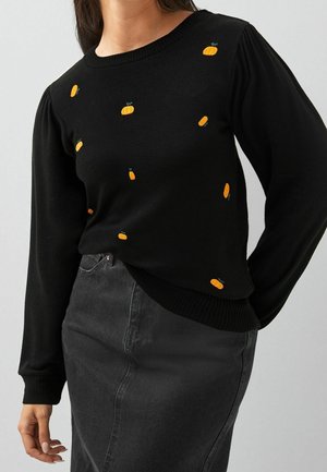 Pull noir à manches longues avec de petites citrouilles orange brodées, porté avec une jupe en jean noire taille haute.