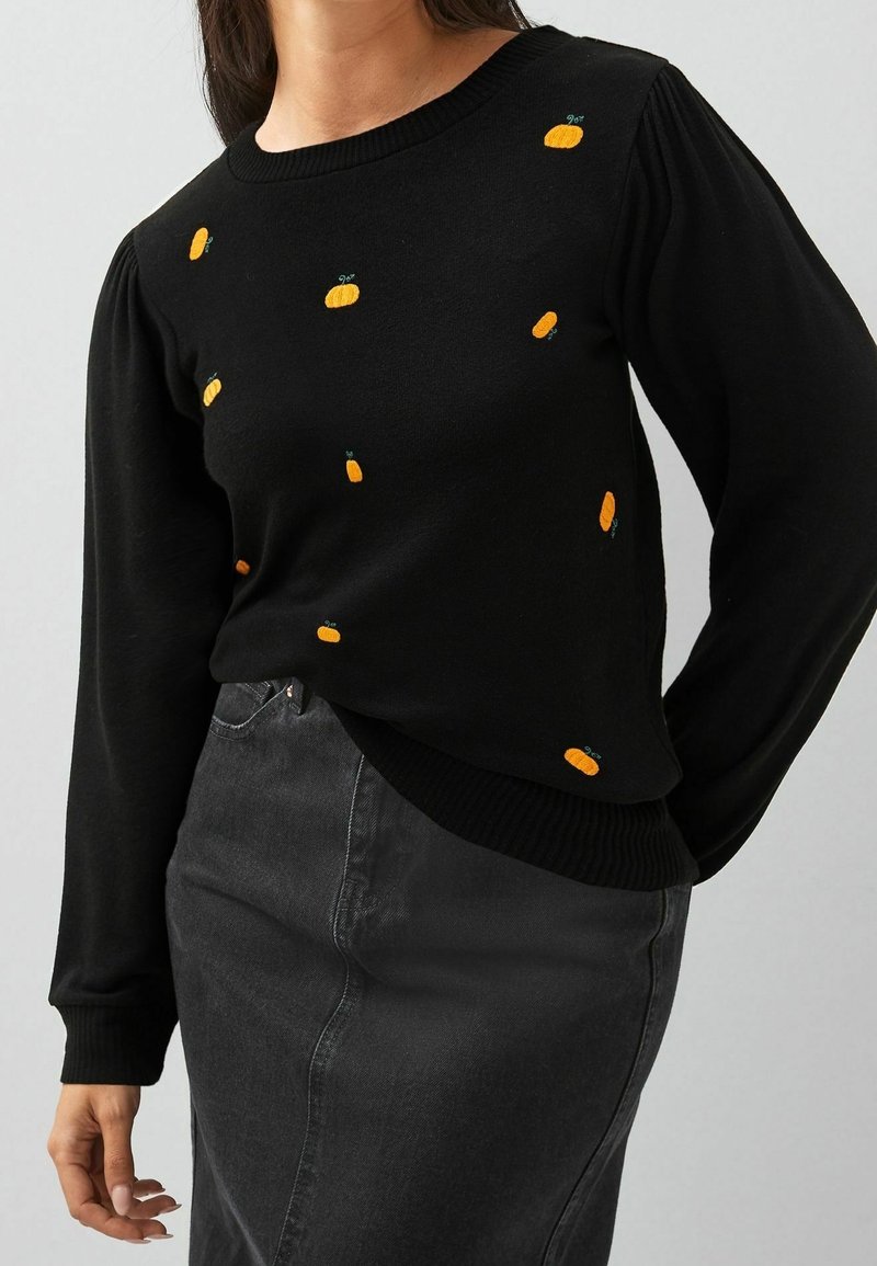 Pull noir à manches longues avec de petites citrouilles orange brodées, porté avec une jupe en jean noire taille haute.