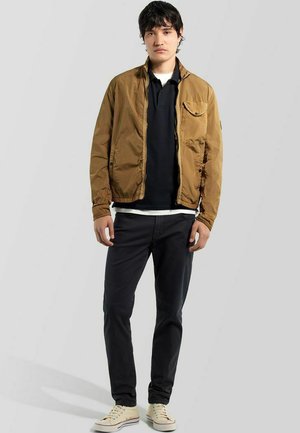 Junger Mann steht und trägt eine braune Jacke über einem schwarzen Poloshirt, dunkle Hosen und beige Turnschuhe vor einem schlichten hellen Hintergrund.