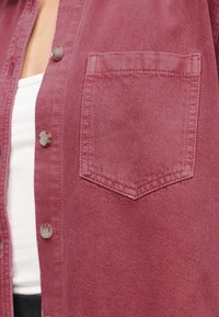 Κοντινό πλάνο ενός ατόμου που φοράει ένα ξεθωριασμένο κόκκινο τζιν πουκάμισο με ανοιχτά κουμπιά πάνω από ένα λευκό τοπ, εμφανίζοντας την τσέπη και τα κουμπιά του πουκαμίσου.