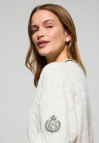 Suéter blanco de punto con patrón de cable texturizado y logo bordado en la manga izquierda. Cuello redondo y corte relajado.