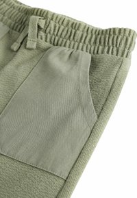 Hellgrüne Jogginghose aus leichter Olive mit elastischem Bund, strukturiertem Stoff und einer glatten Pattentasche an der Seite.