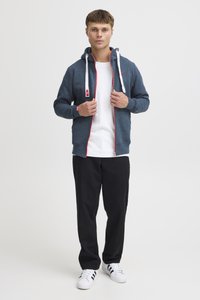 Marineblauwe hoodie met ritssluiting, rode accenten en witte trekkoorden, gedragen over een wit T-shirt, gecombineerd met zwarte broek en witte sneakers.