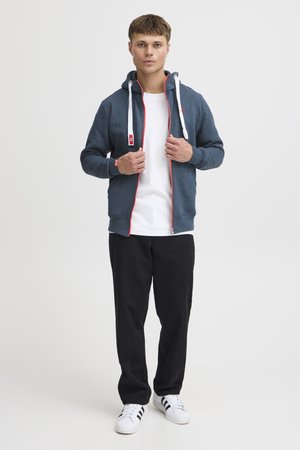 Felpa zipata blu scuro con lacci bianchi, dettagli rossi, indossata sopra una maglietta bianca. Pantaloni neri e sneakers bianche con righe nere.