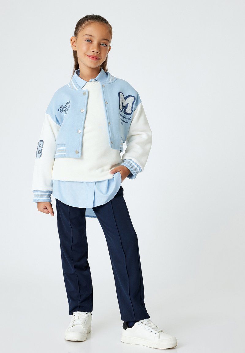 Varsityjack in lichtblauw en wit met geborduurde details, gelaagd over een lichtblauwe shirt en marineblauwe broek. Witte sneakers maken de look compleet.
