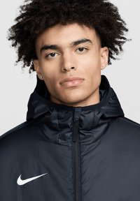 Schwarze Nike-Jacke mit Kapuze, ausgestattet mit einem durchgehenden Reißverschluss, weichem Stoff und einem auffälligen weißen Logo auf der linken Brust.