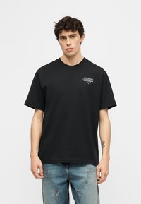 Νέος με σγουρά μαλλιά, που φοράει μαύρο t-shirt Puma με μικρό άσπρο λογότυπο στο στήθος και μπλε τζιν, στέκεται μπροστά σε ένα απλό φόντο.