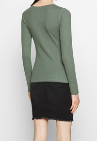 Ribbad långärmad topp i dämpad grön, ihop med en svart denimkjol med fransat nederkant. Formsydd design med rund halsringning.