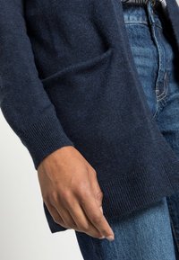 Marineblaue Strickjacke mit langen Ärmeln, gerippten Bündchen und einer Taschenpartie; kombiniert mit blauen Denim-Jeans, die eine strukturierte Oberfläche aufweisen.