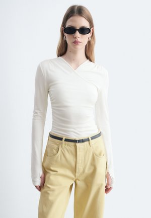 Jeune femme portant des lunettes de soleil ovales noires, un haut blanc ajusté à manches longues, un pantalon beige et une ceinture noire, debout devant un fond blanc.