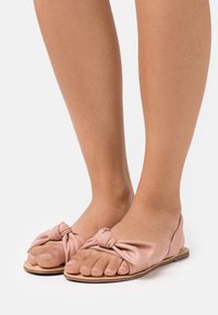 Sandali piatti di colore rosa chiaro realizzati in morbida pelle, con un dettaglio a fiocco annodato nella parte anteriore. Il design open-toe mette in mostra le unghie dei piedi ben curate.