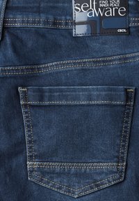 Vaqueros de mezclilla en azul oscuro con un solo bolsillo trasero. Presentan costuras visibles en beige claro. Etiqueta de la marca en la cinturilla.