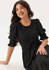 Abito nero con maniche corte a palloncino e corpetto arricciato. Texture morbida, lunghezza al ginocchio e silhouette rilassata. Presenta dettagli arricciati.