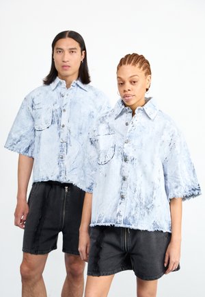 Zwei Models tragen hellblaue, übergroße Jeanshemden mit kurzen Ärmeln und fransigen Kanten. Die Hemden haben zwei vordere Taschen und Knopfverschlüsse.