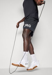 Weiße Sportschuhe mit strukturierten Sohlen, schwarzem Logo, grauen Shorts mit "ORG"-Aufdruck und weißen Knöchelsocken, während ein Springseil gehalten wird.