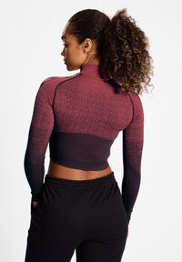 Langärmliges, gekürztes Sportoberteil in einem Farbverlauf von Rot zu Schwarz, mit einem strukturierten Muster und enganliegendem Design. Kombiniert mit lockeren schwarzen Hosen.