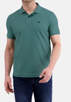 Mann trägt ein türkisfarbenes kurzärmeliges Poloshirt mit Zwei-Knopf-Leiste und dunkelblaue Hose, steht vor einem schlichten Hintergrund.