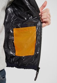 Schwarze, glänzende, wasserfeste Jacke mit innerem orangen Netzfach. Verfügt über einen Reißverschluss und einen elastischen Saum, der eine glatte, strukturierte Oberfläche zeigt.