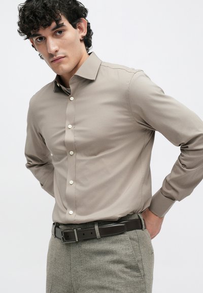 Beige, long-sleeve button-up shirt made of smooth fabric. Features a classic collar, front buttons, and is paired with textured gray trousers.