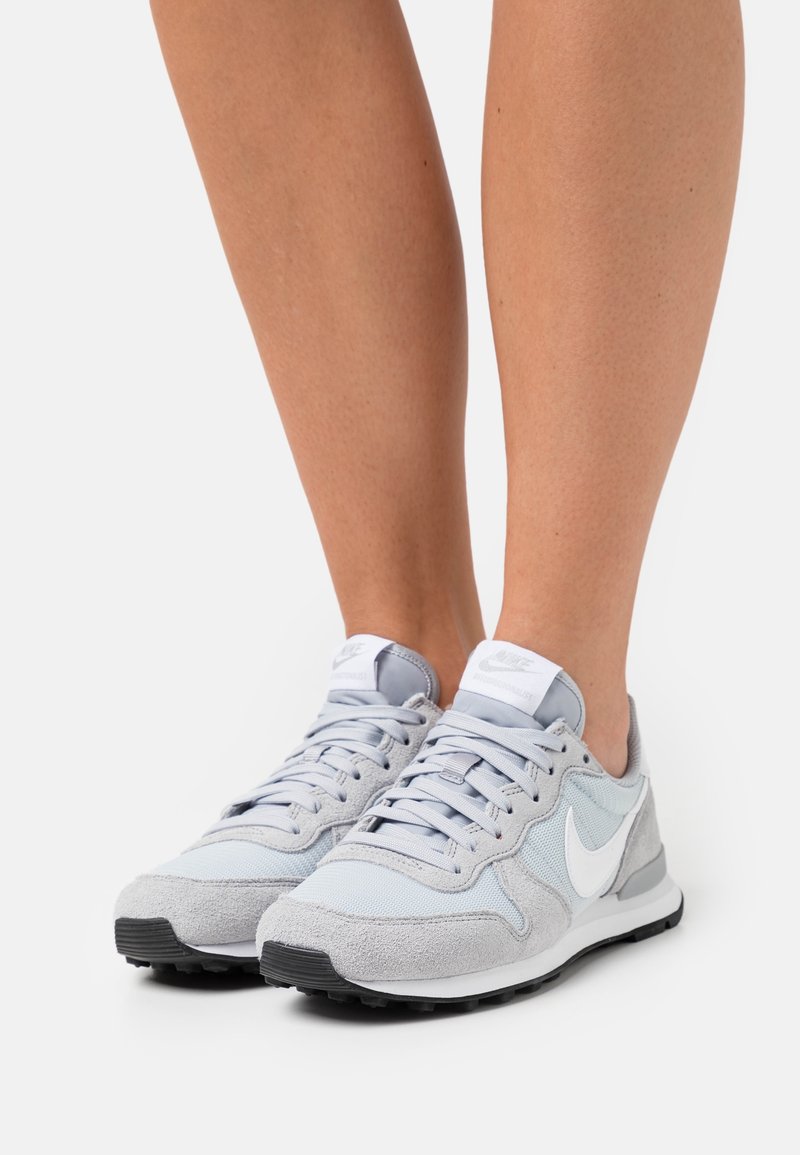 Personne portant des baskets Nike gris clair et blanc avec des semelles noires, debout sur une surface blanche.