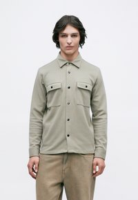 Νέος άνδρας φορώντας ένα ανοιχτό γκρι πουκάμισο με δύο κουμπωτές τσέπες στο στήθος και μπεζ παντελόνι, που στέκεται μπροστά σε ένα απλό λευκό φόντο.