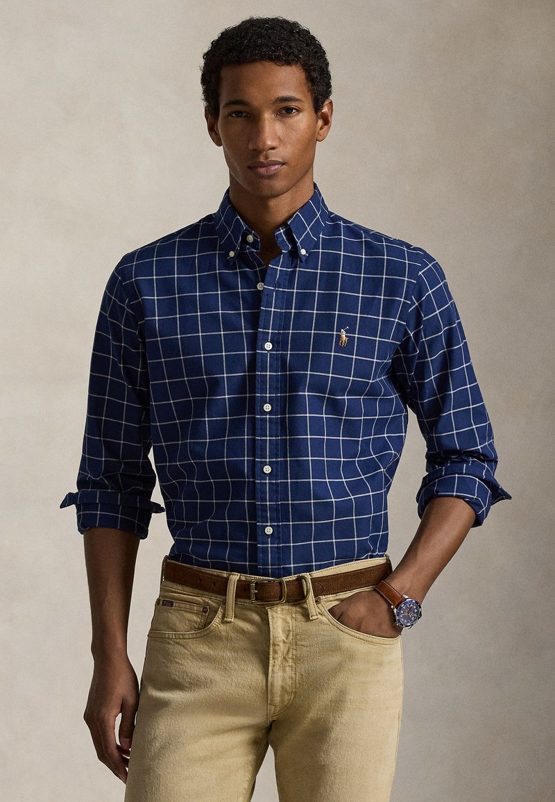 Navy blue checked shirt with white grid pattern, button-down collar, and rolled sleeves, paired with light beige jeans and a brown belt.