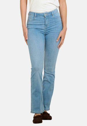 Kobieta ubrana w jasnoniebieskie, wysokowalne jeansy o prostych nogawkach z pięcioma kieszeniami oraz brązowe mokasyny, stojąca na białym tle.