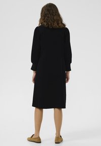Black corduroy dress with long puff sleeves, knee-length, featuring a fitted bodice and flowing skirt, paired with light brown slip-on shoes.