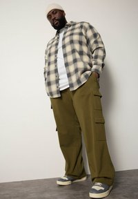 Gematigde flanellen shirt in beige en grijs over een wit T-shirt, gecombineerd met olijfgroene cargobroeken en grijze sneakers.