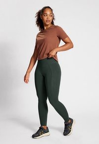 Donkergroene geribbelde leggings met een hoge tailleband, gecombineerd met een bruin shirt met korte mouwen. Model draagt zwarte en gele sportschoenen.