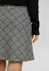 Acercamiento de una falda gris con un sutil patrón de cuadros oscuros, combinada con una blusa negra de manga larga de punto, mostrando una mano y parte de las piernas.