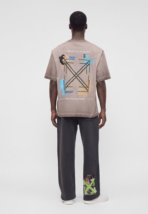 Une personne est debout, dos à nous, portant un t-shirt beige avec des motifs graphiques et un pantalon gris foncé avec un imprimé vert et marron près de la cheville gauche.