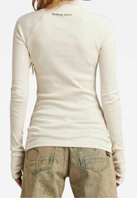 Top de manga larga color crema con un cuello alto. Presenta el logo de G-Star Raw en la parte posterior, combinado con pantalones de mezclilla de tono verdoso.