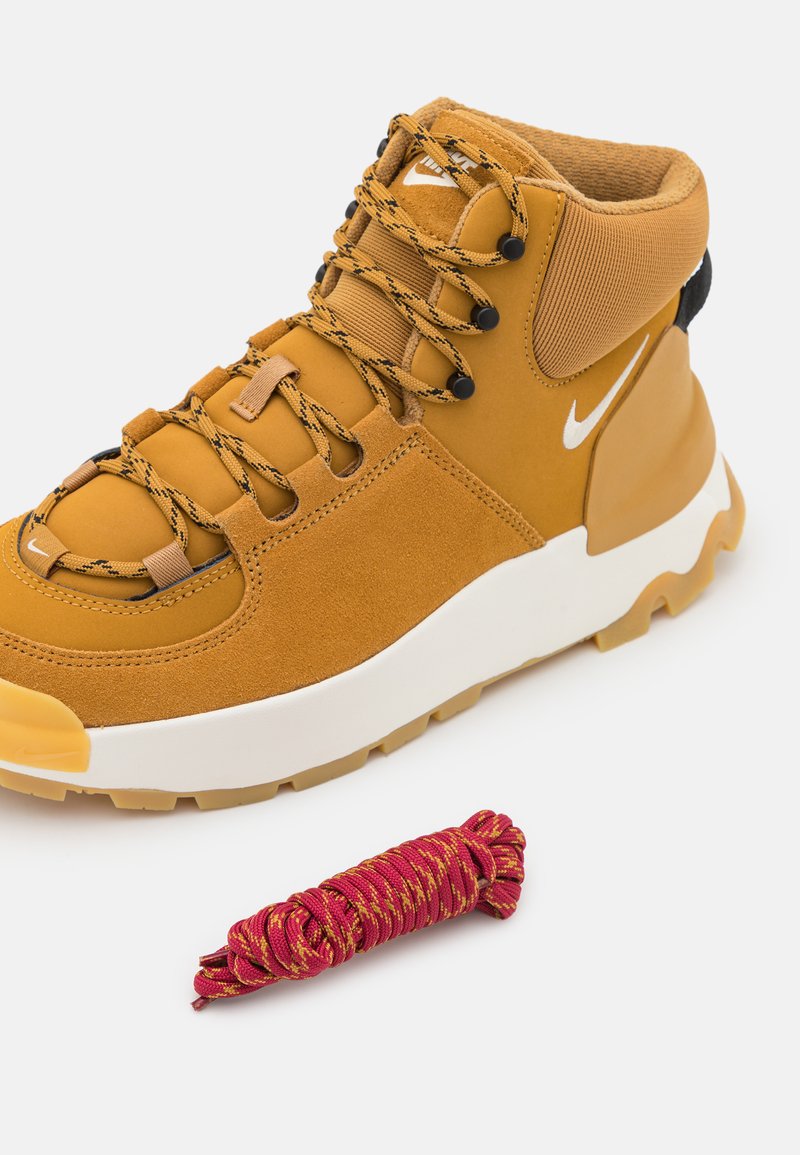 Nike-boots in mosterdgele suede en synthetisch materiaal met zwarte accenten, voorzien van zwarte veters en een witte middenzool. Inclusief een rode paracord.