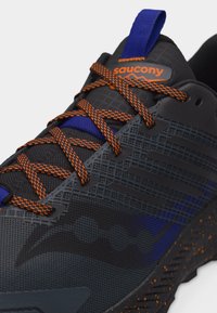 Μαύρο και μπλε αθλητικό παπούτσι Saucony με κορδόνια και σόλα πορτοκαλί με στίγματα, κοντινή λήψη που δείχνει υφασμάτινο δίχτυ με υφή και το λογότυπο της μάρκας στη γλώσσα.