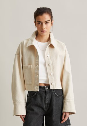 Jeune femme portant une veste beige courte boutonnée par-dessus un crop top blanc et un short en denim noir taille haute, debout devant un fond neutre.
