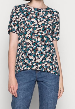 Woman wearing a short-sleeve, crew-neck floral shirt with white, beige, and teal flowers paired with blue jeans.