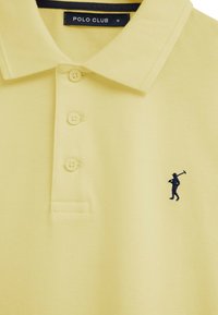 Polo jaune à trois boutons, avec un col, et un logo brodé bleu marine représentant un golfeur portant un club sur le côté gauche de la poitrine.