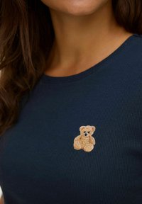 Marineblå ribbet striktrøje med en lille, broderet brun teddybjørn på venstre bryst og rund halsudskæring.