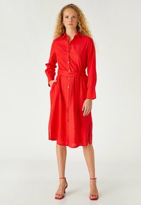 Vestido rojo abotonado, hecho de una tela ligera, con mangas largas, cintura con cinturón y bolsillos laterales para mayor funcionalidad.