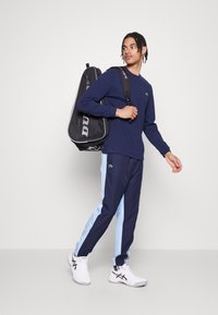 Ναυτικό πουκάμισο μακρυμάνικο σε συνδυασμό με σορτς αθλητικού στιλ σε ναυτικό και ανοιχτό μπλε χρώμα. Λευκά αθλητικά παπούτσια με μαύρες λεπτομέρειες, κρατάει μια μαύρη αθλητική τσάντα.