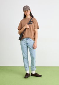 Camiseta de algodón marrón con un logo negro, jeans ajustados de mezclilla azul claro, calcetines blancos y zapatos marrones oscuros. Una gorra gris completa el atuendo.