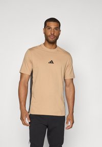 Beige sporty t-shirt med korte ærmer, rund halsudskæring, sort logo på brystet og sorte sidelister. Blødt stof, tætsiddende pasform.