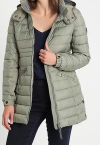 Chaqueta de plumas verde oliva con textura acolchada, cremallera frontal y capucha desmontable con botones a presión. Cuenta con bolsillos laterales y forro gris en contraste.