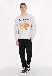 Hanorac gri cu text grafic mixat în culori vibrante, asortat cu pantaloni negri și adidași negri cu accente roșii. Fundal simplu.